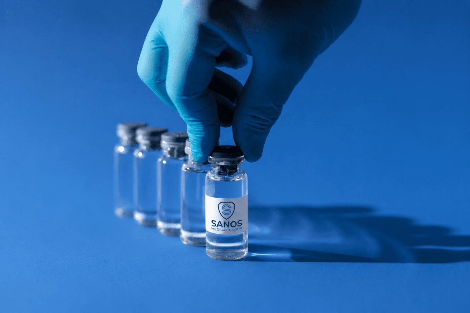 Una mano con guante seleccionando un vial clínico con la etiqueta de Sanos Medical Group de una fila de viales